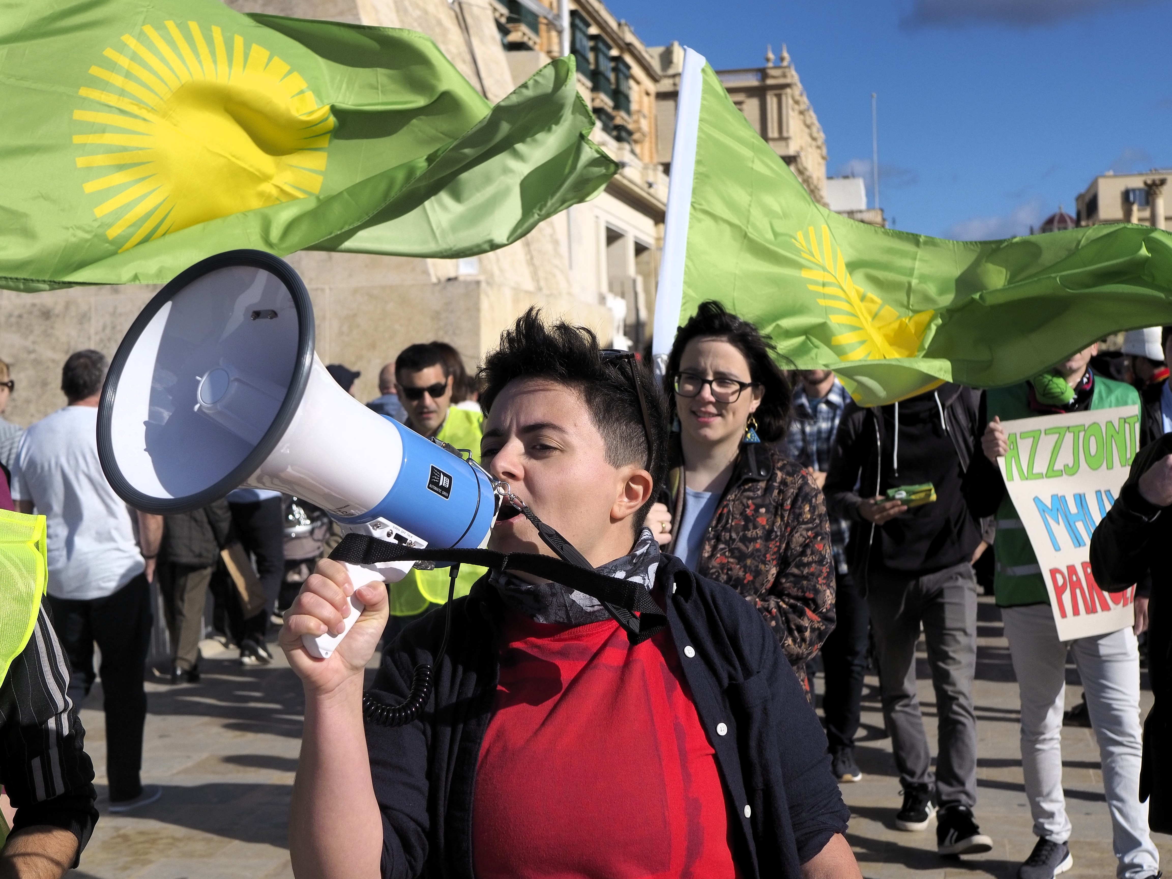 Mina Tolu at the foreground of the photo holds a white and blue megaphone that they talk into. Behind them are other protestors holding green and yellow flags and posters.